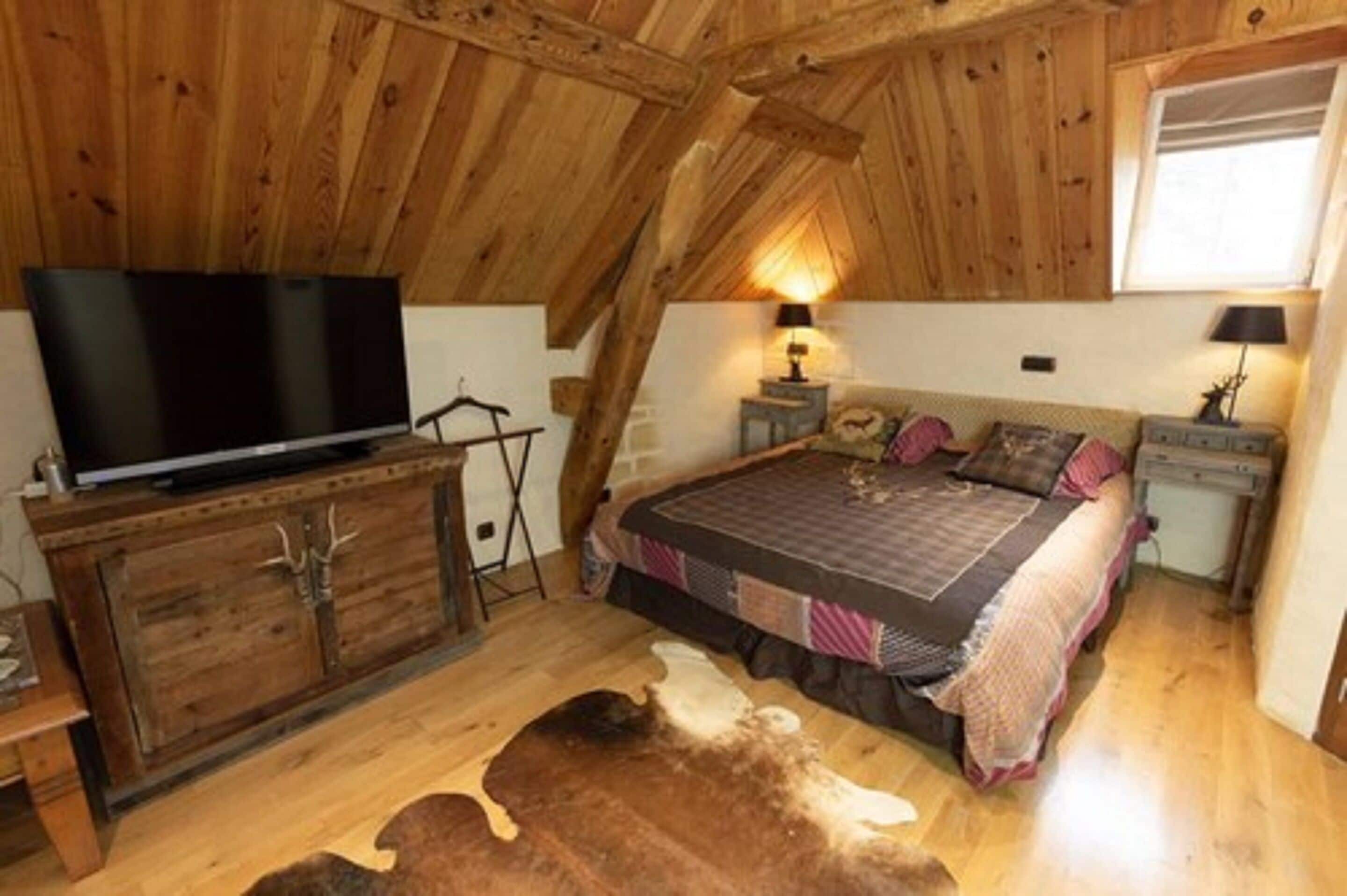 A cozy attic bedroom with a wooden ceiling, a large bed, a TV on a wooden cabinet, and a cowhide rug.