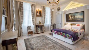 A bedroom with a large bed, a chandelier, a painting, and a desk.