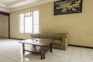 A room with a beige sofa, a wooden coffee table, a painting of birds on the wall, and a window with blinds. - Hotel O Padjadjaran 1 (Tasikmalaya)