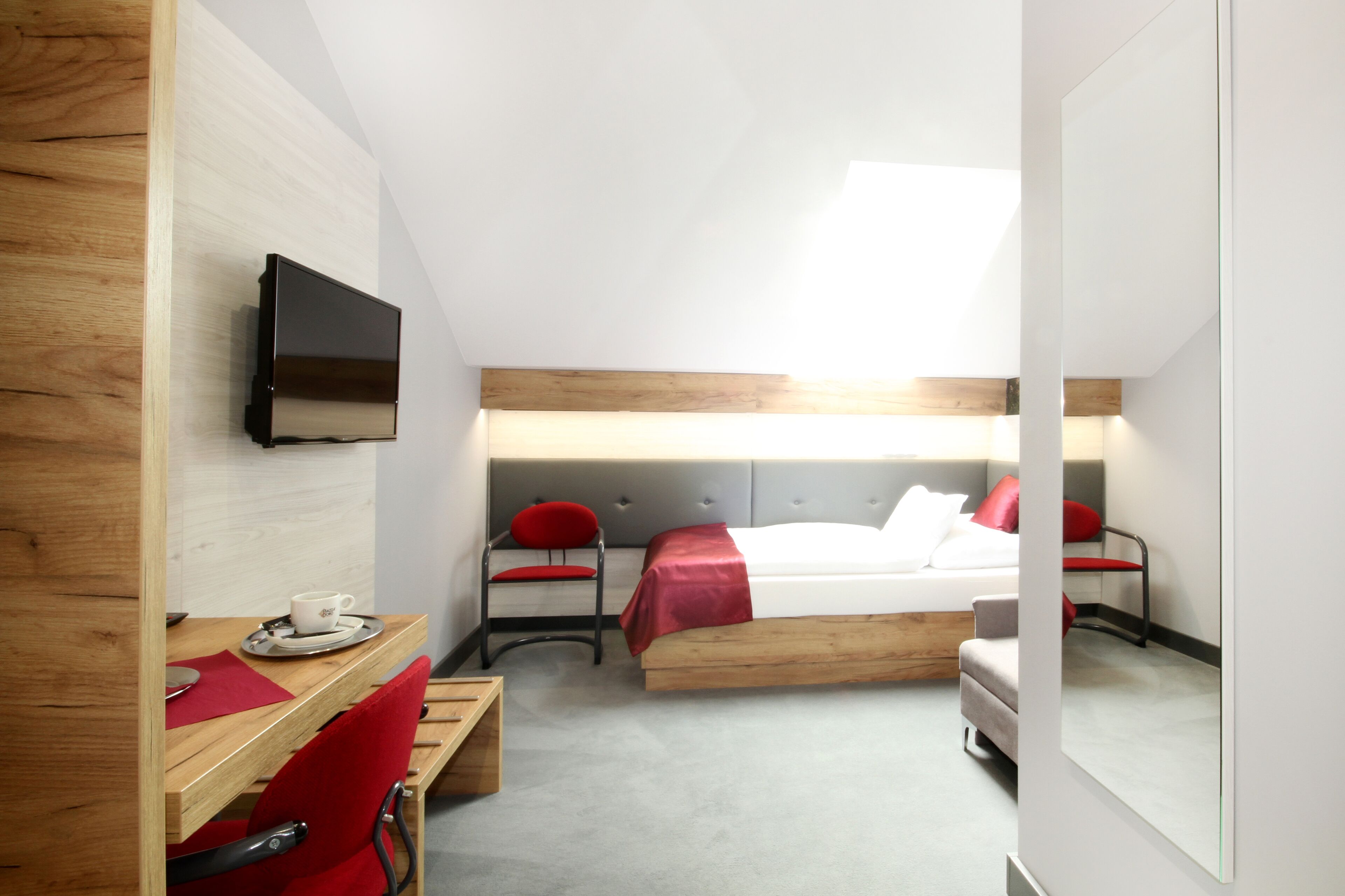Habitación de hotel moderna con un escritorio de madera, un televisor montado en la pared, una cama con colcha roja y blanca, y una pequeña zona de estar con un sillón rojo.