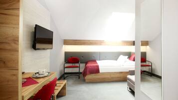 Habitación de hotel moderna con un escritorio de madera, un televisor montado en la pared, una cama con colcha roja y blanca, y una pequeña zona de estar con un sillón rojo.