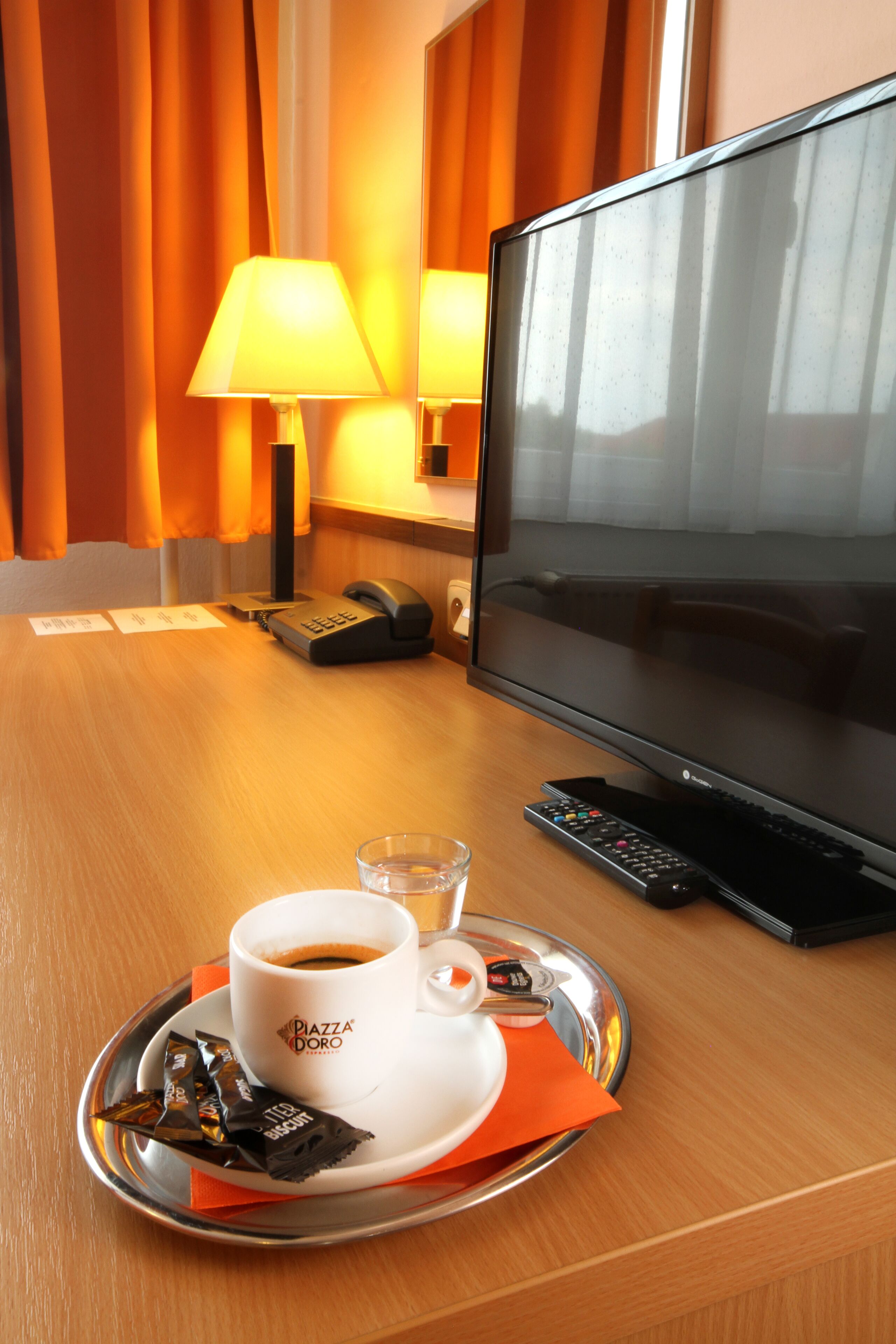 Habitación de hotel con una mesa de madera, un televisor, una lámpara, una taza de café, un vaso de agua y una bandeja con galletas de biscotti.