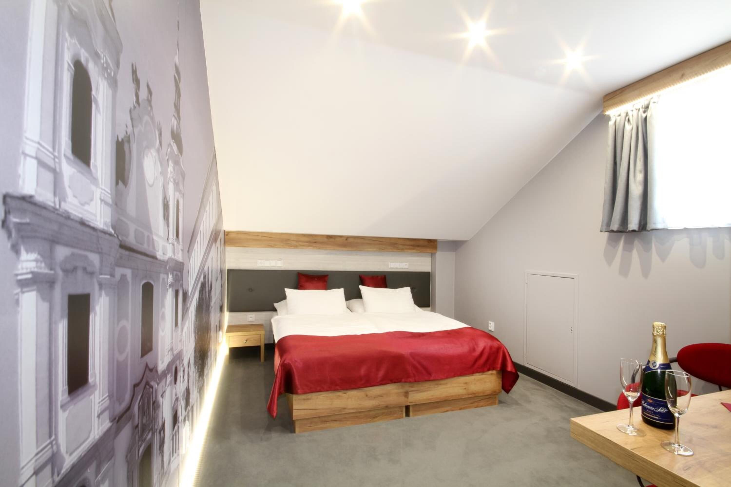 Un dormitorio con cama, mesita de noche, cabecero de madera, colcha roja, un mural en la pared, una ventana con cortinas y una botella de champán sobre una mesa.