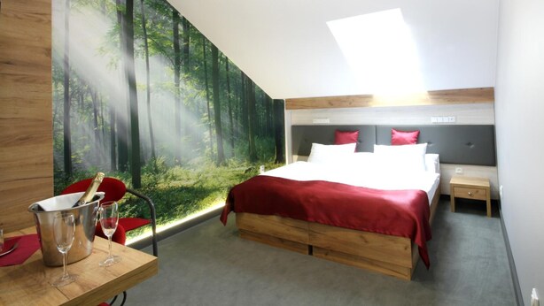 A bedroom with a bed, a nightstand, a wooden desk with a bottle and glasses, and a forest mural on the wall. - Hotel Strážnice (Straznice)