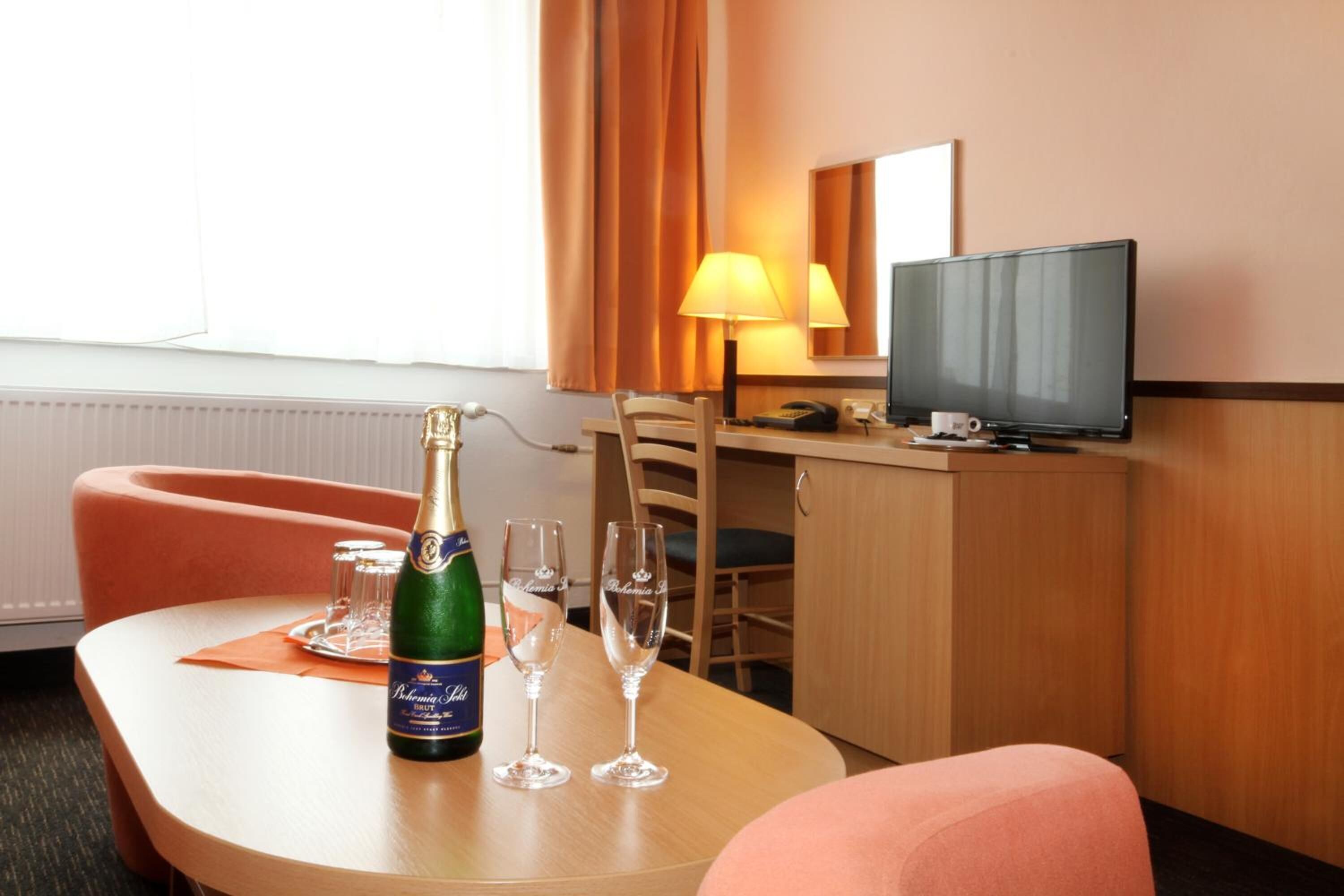 A hotel room with a wooden desk, a television, a bottle of champagne, and two glasses.