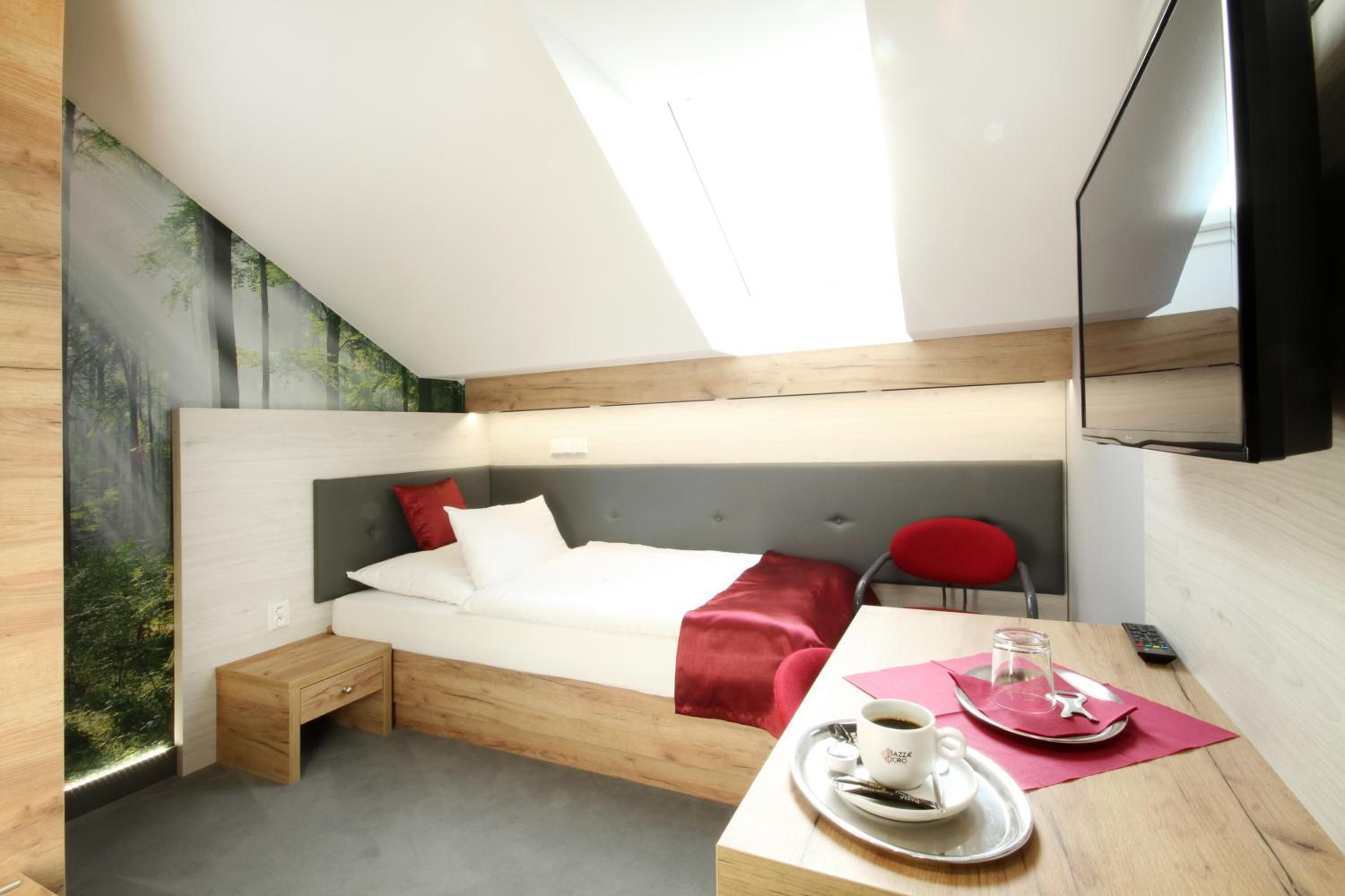 A modern hotel room with a bed, a small desk with a cup of coffee, a red chair, and a TV.
