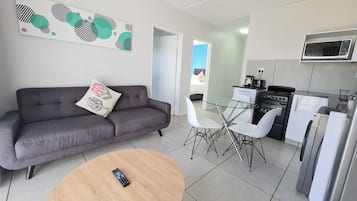 En moderne stue med en grå sofa, et rundt træbord og et spisebord i glas med hvide stole. Et køkken med komfur, mikrobølgeovn og køleskab er synligt i baggrunden.