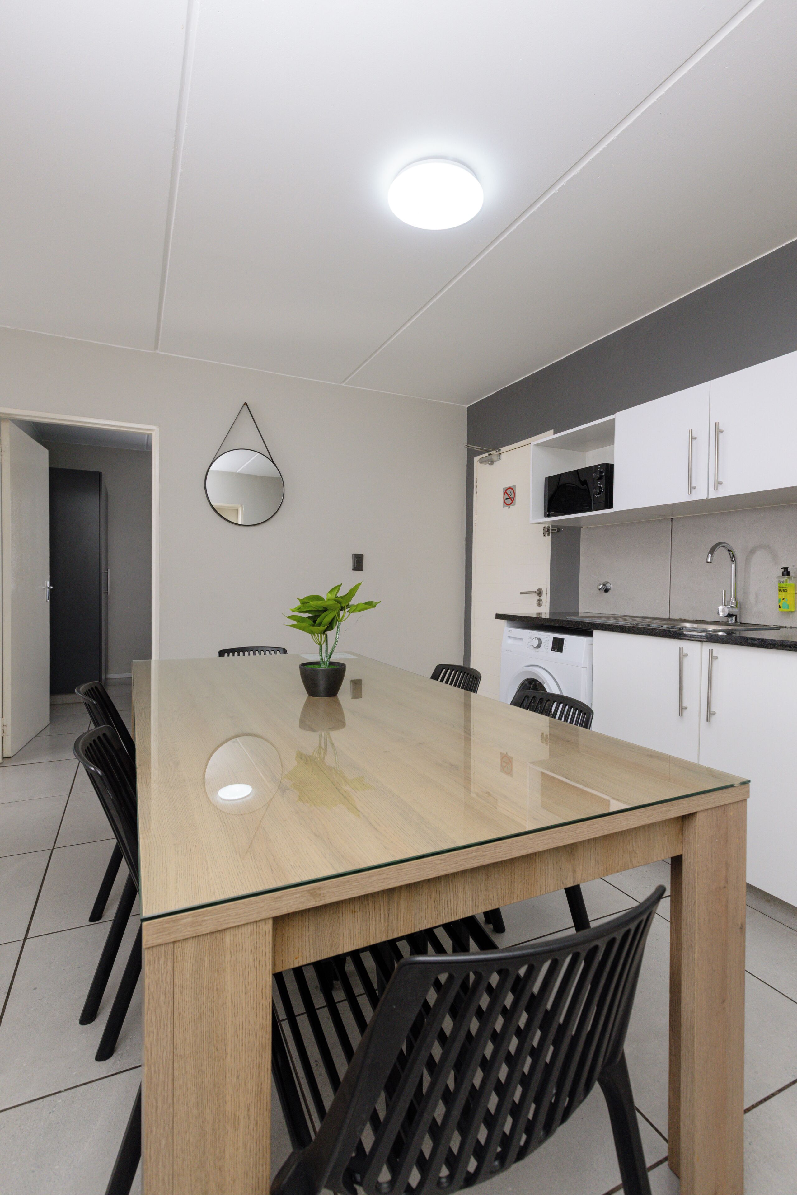 A modern kitchen with a glass-top dining table, black chairs, a potted plant, and white cabinets.