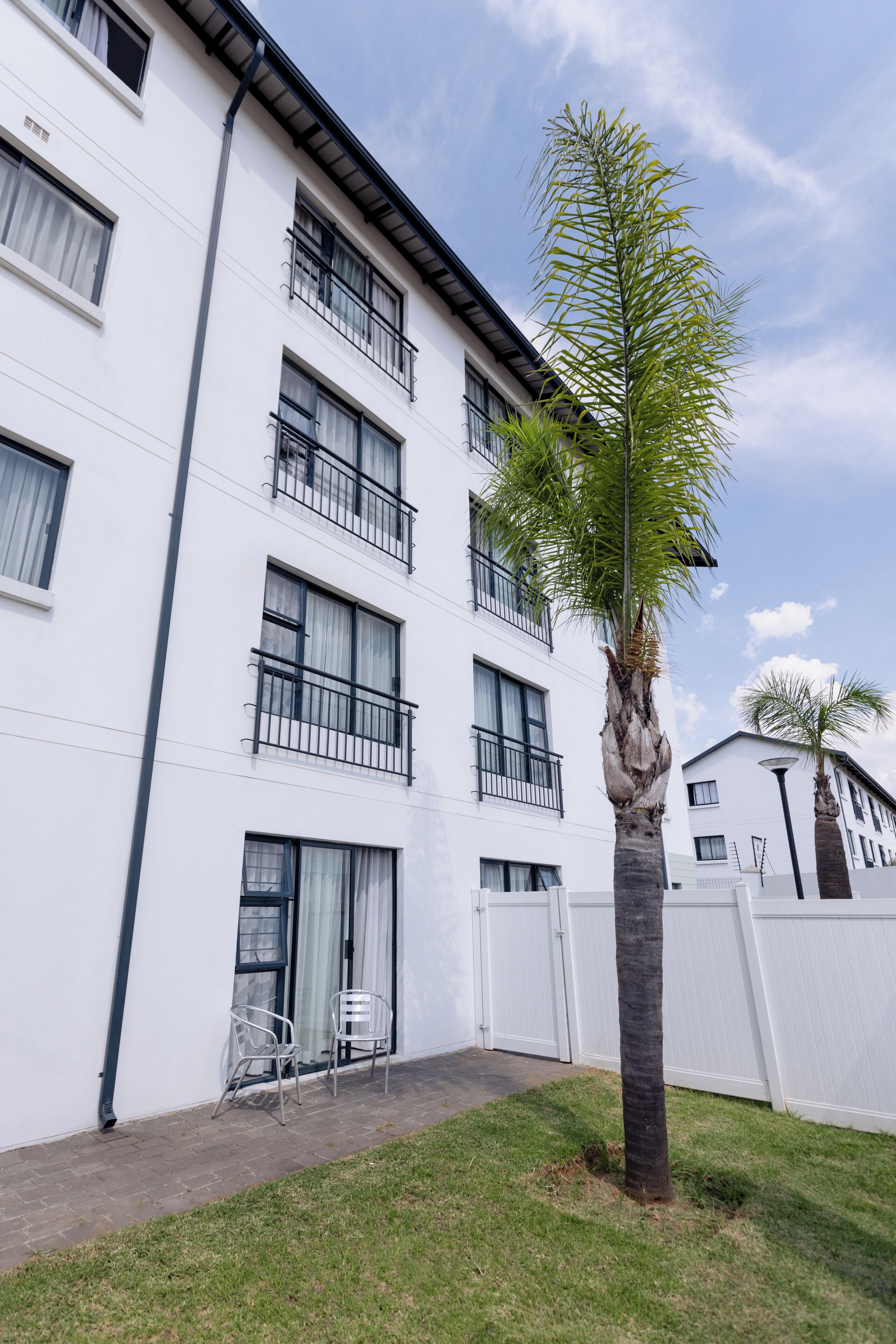 A white multi-story building with balconies, a palm tree, and a white fence.
