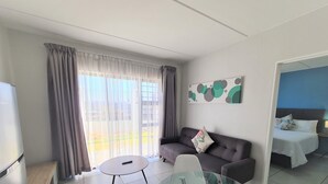 A modern hotel room with a sofa, a glass dining table, a TV, and a bed with a blue wall and a decorative pillow. - MINT Express Sandton View (Sandton)