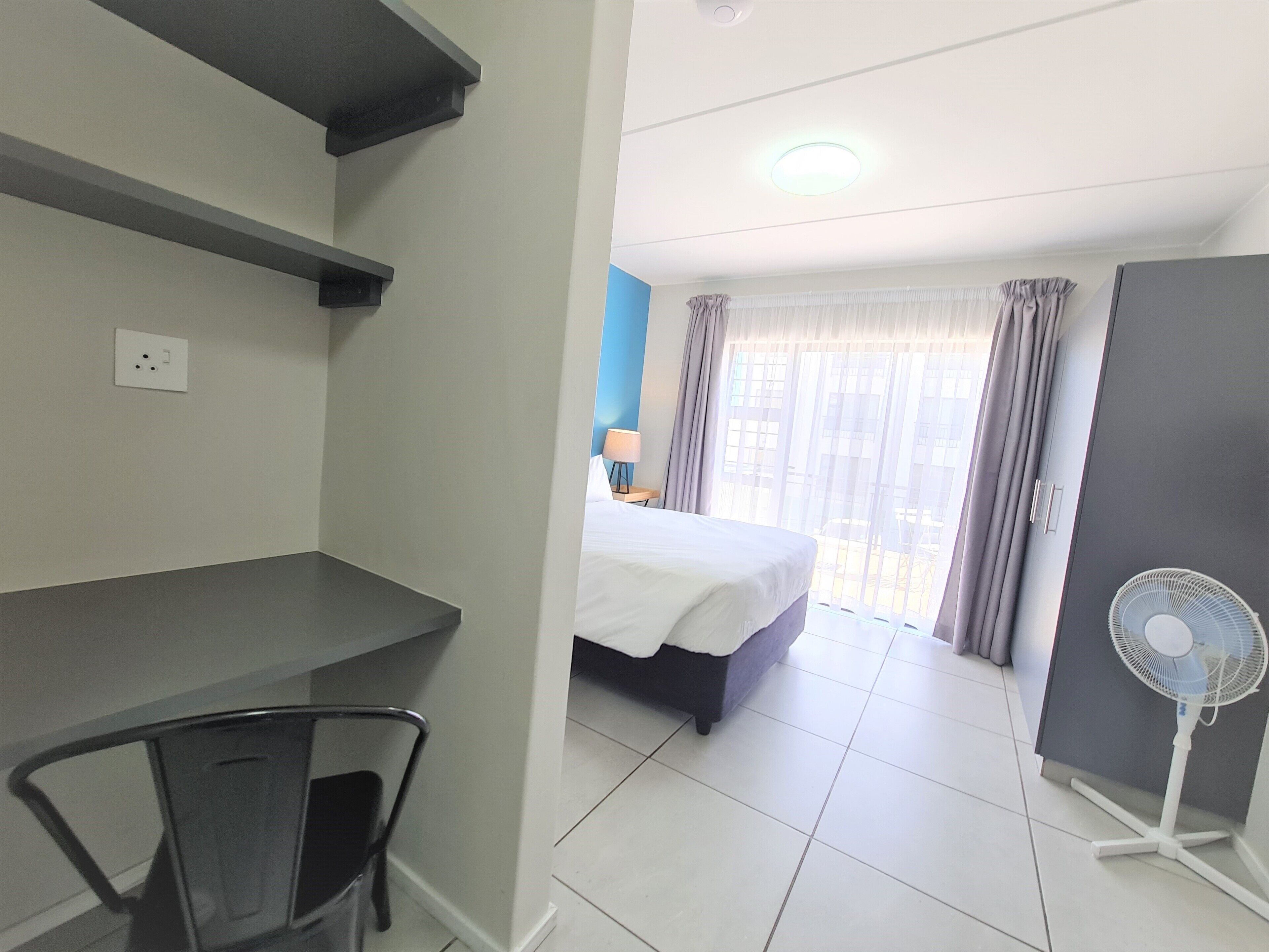 Una habitación de hotel con una cama, un escritorio, una silla, un ventilador de pie y una ventana con cortinas.