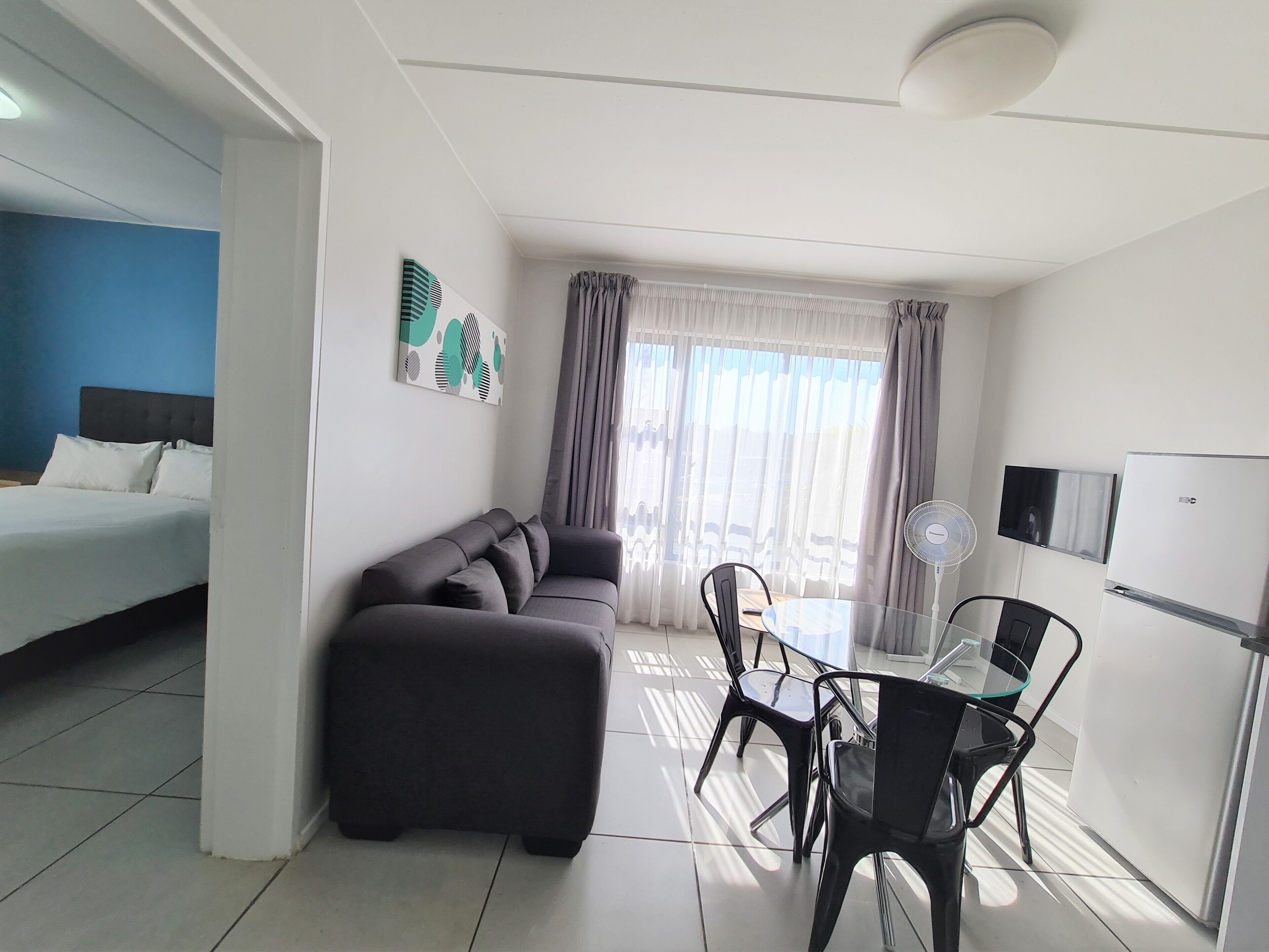A modern hotel room with a bed, sofa, dining table, chairs, and a refrigerator.