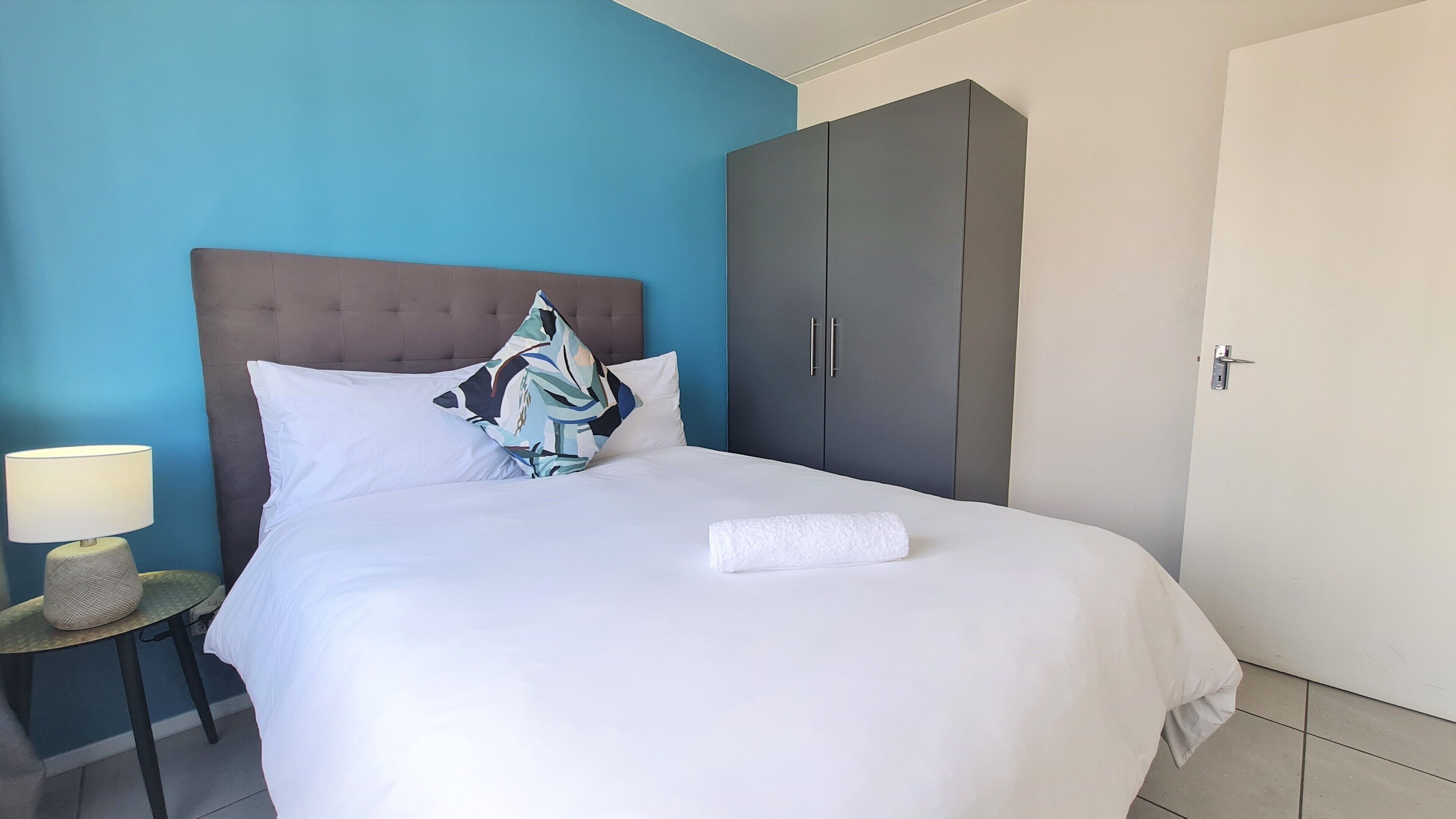 Una cama bien tendida con sábanas blancas y una toalla blanca, pared azul, armario gris y mesita de noche con lámpara.