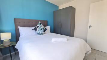Una cama bien tendida con sábanas blancas y una toalla blanca, pared azul, armario gris y mesita de noche con lámpara.
