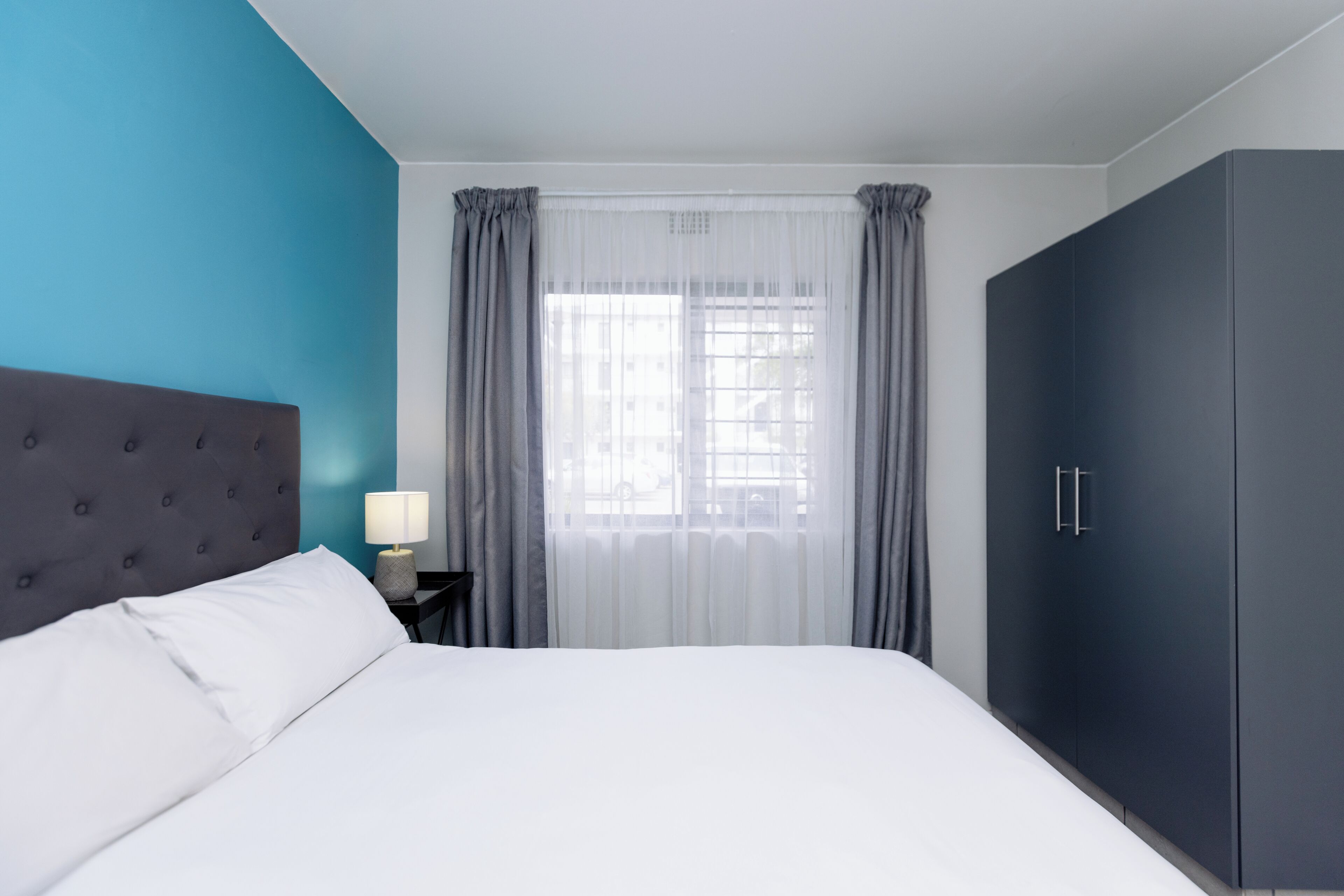A bedroom with a bed, a nightstand, a lamp, a window with curtains, and a wardrobe.