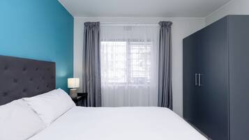 A bedroom with a bed, a nightstand, a lamp, a window with curtains, and a wardrobe.