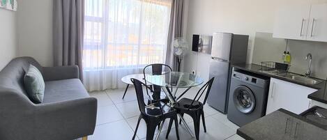 Un espacio habitable funcional con sofá, mesa de comedor, sillas, lavadora y cocina equipada con armarios y fregadero.