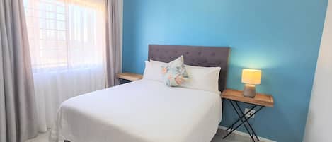 A neatly made bed with white linens, a blue wall, and a window with curtains.