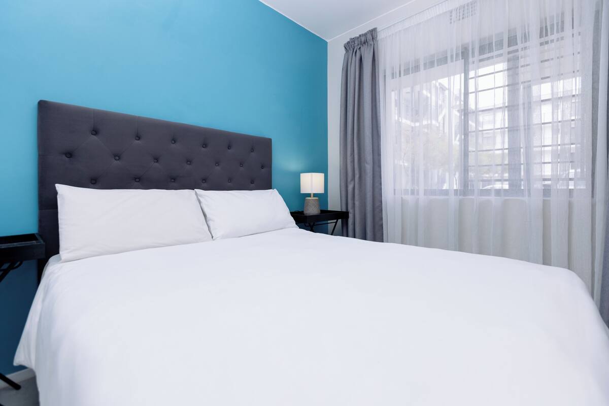 A bed with a tufted headboard, a bedside table with a lamp, and a window with sheer curtains.