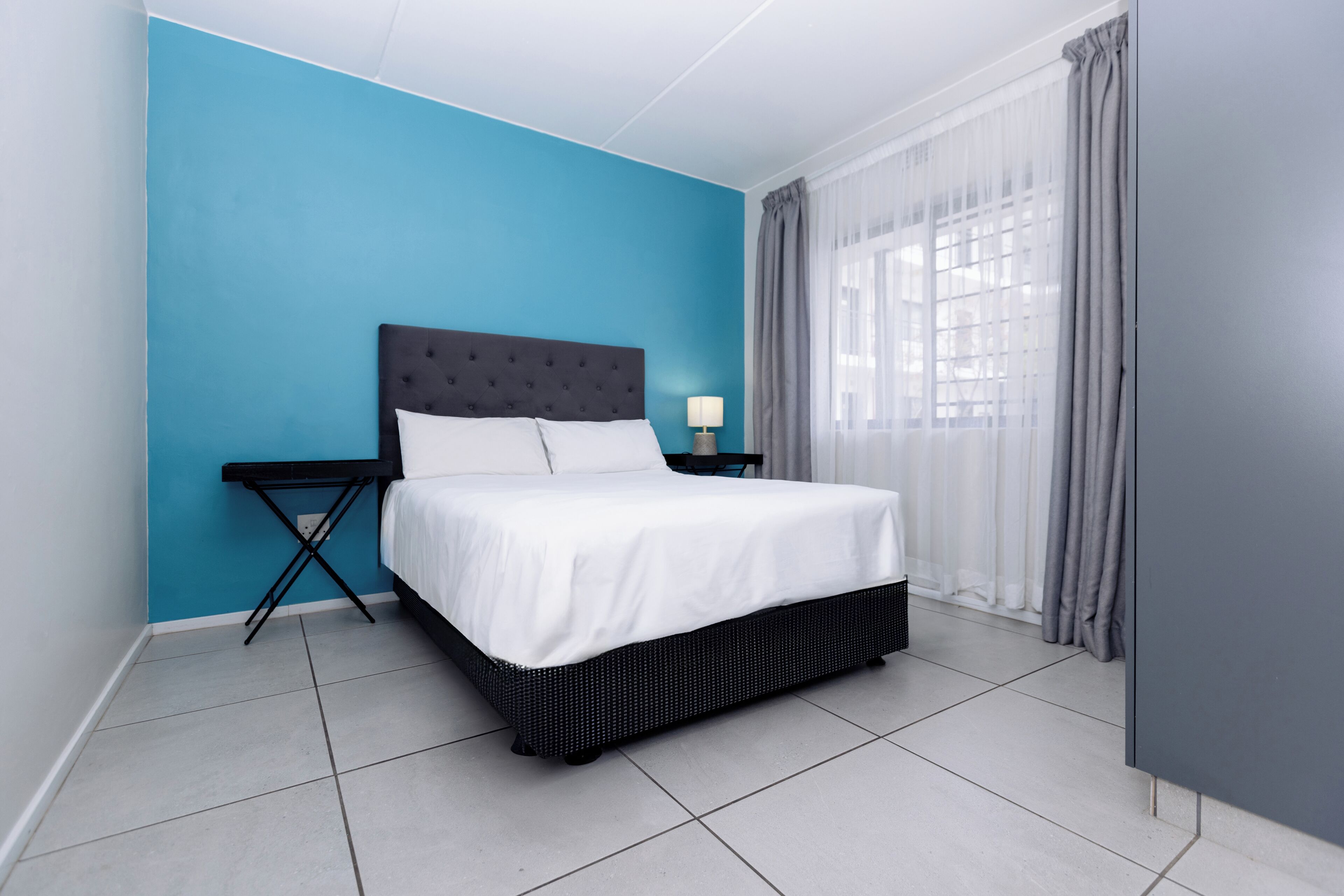 A bed with a white mattress and dark headboard, a small bedside table with a lamp, and a window with grey curtains.