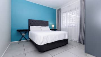 A bed with a white mattress and dark headboard, a small bedside table with a lamp, and a window with grey curtains.
