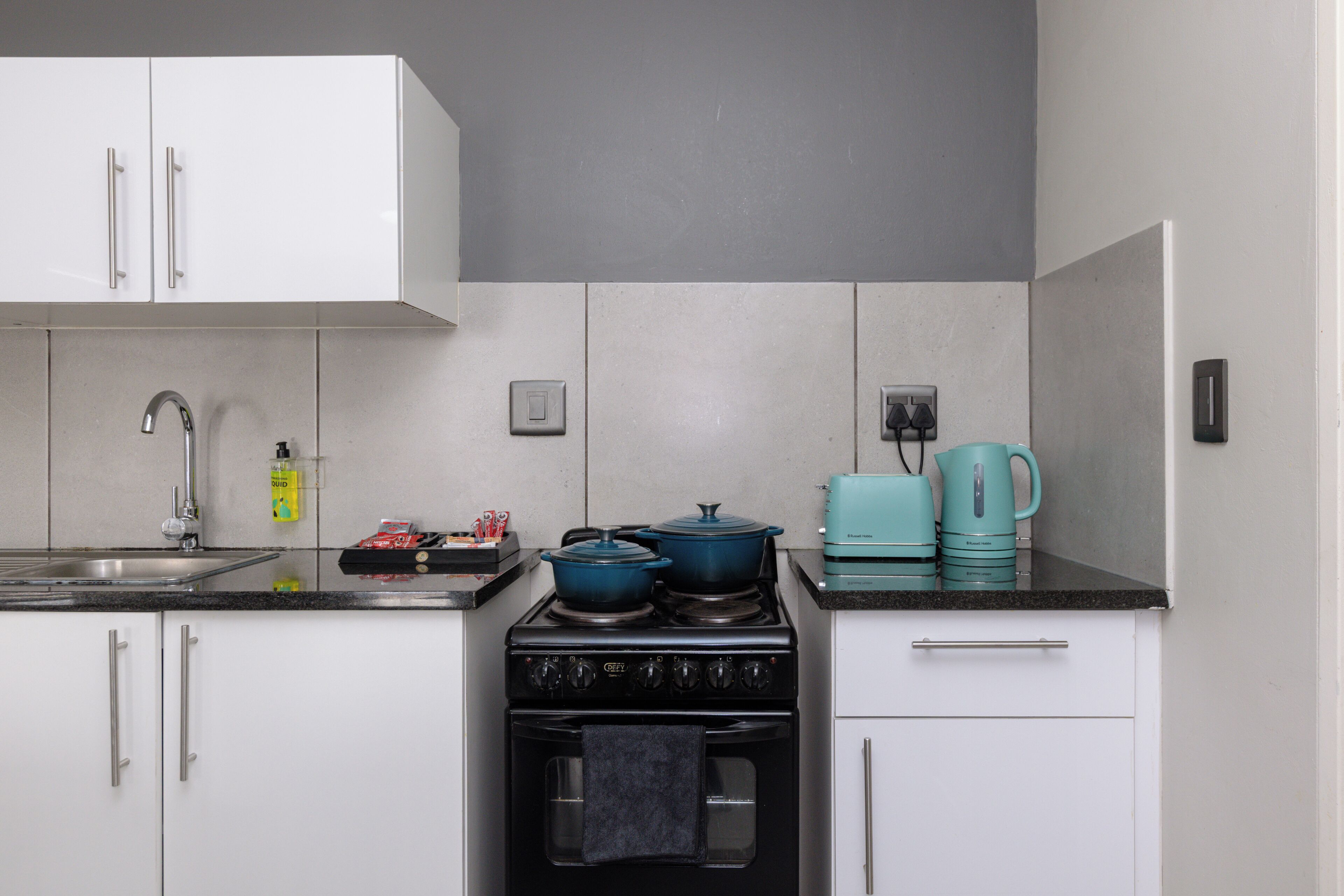 Une cuisine avec des meubles blancs, un poêle noir, un évier et une cafetière turquoise.
