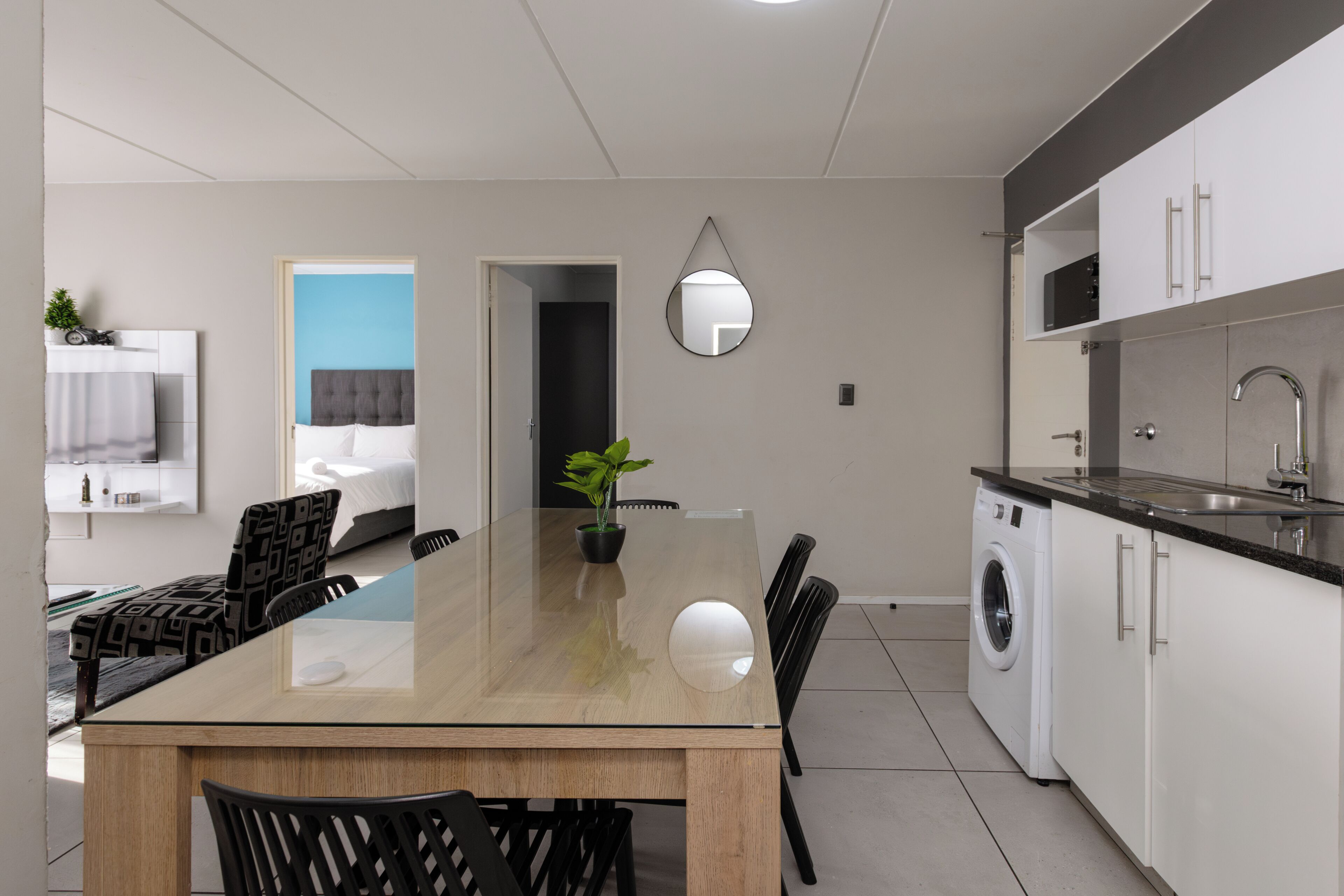 Eine moderne Küche mit Waschmaschine, eine Essecke mit Glastisch und ein Schlafzimmer mit Bett und Fernseher.