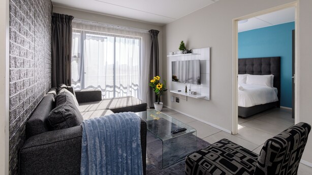 A modern living room with a sofa, a glass coffee table, a TV, and a bed in the adjacent room. - MINT Express Sandton View (Sandton)