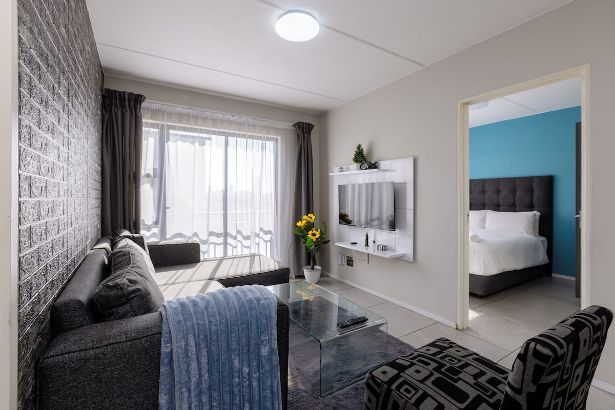 A modern living room with a sofa, a glass coffee table, a TV, and a bed in the adjacent room.