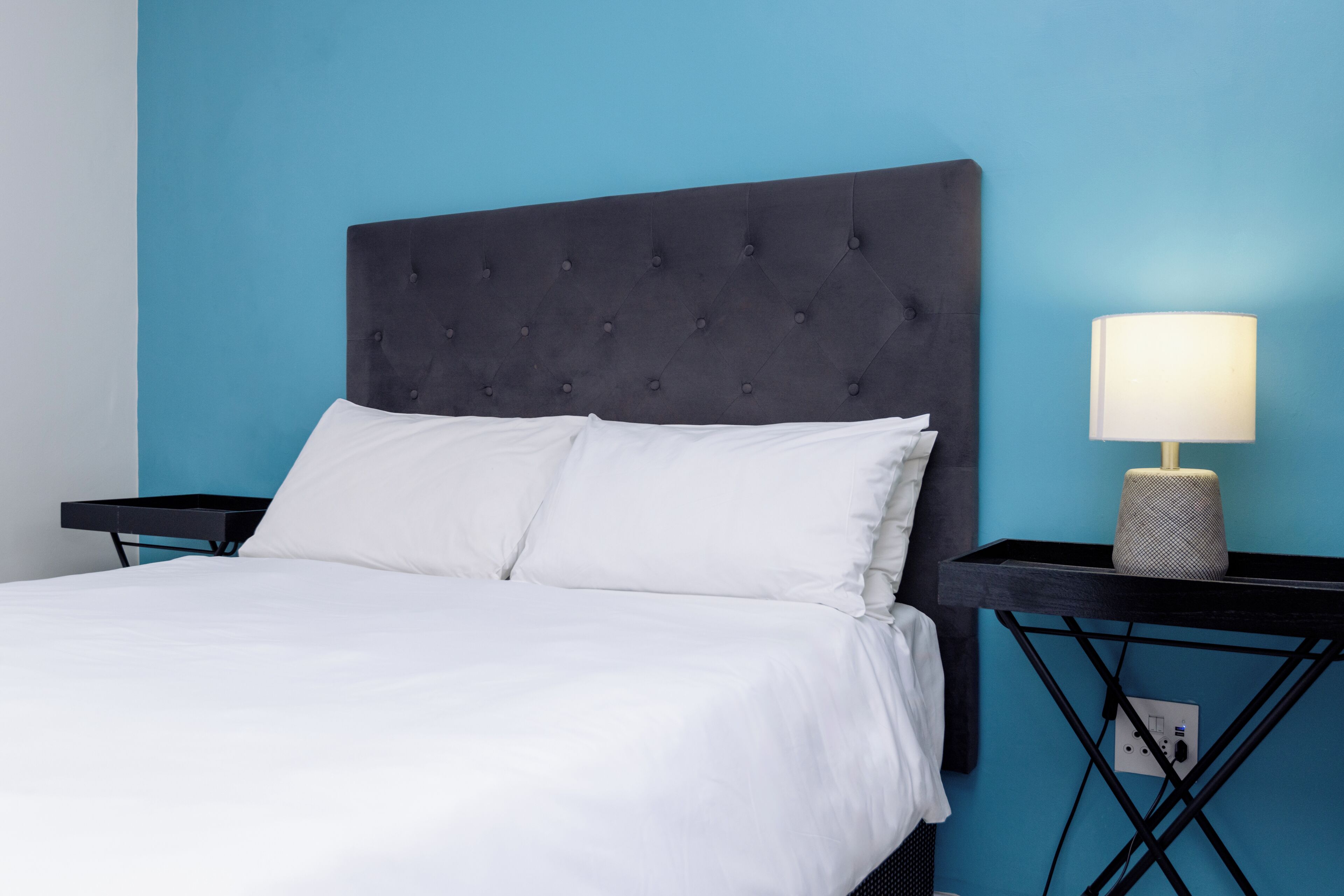 Un lit doté d’une tête de lit capitonnée, de draps blancs et d’une lampe de chevet posée sur une table de nuit noire.
