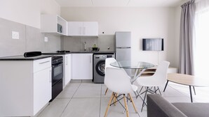 A modern kitchen with white cabinets, a dishwasher, a washing machine, and a dining area with a glass table and white chairs. - MINT Express Sandton View (Sandton)