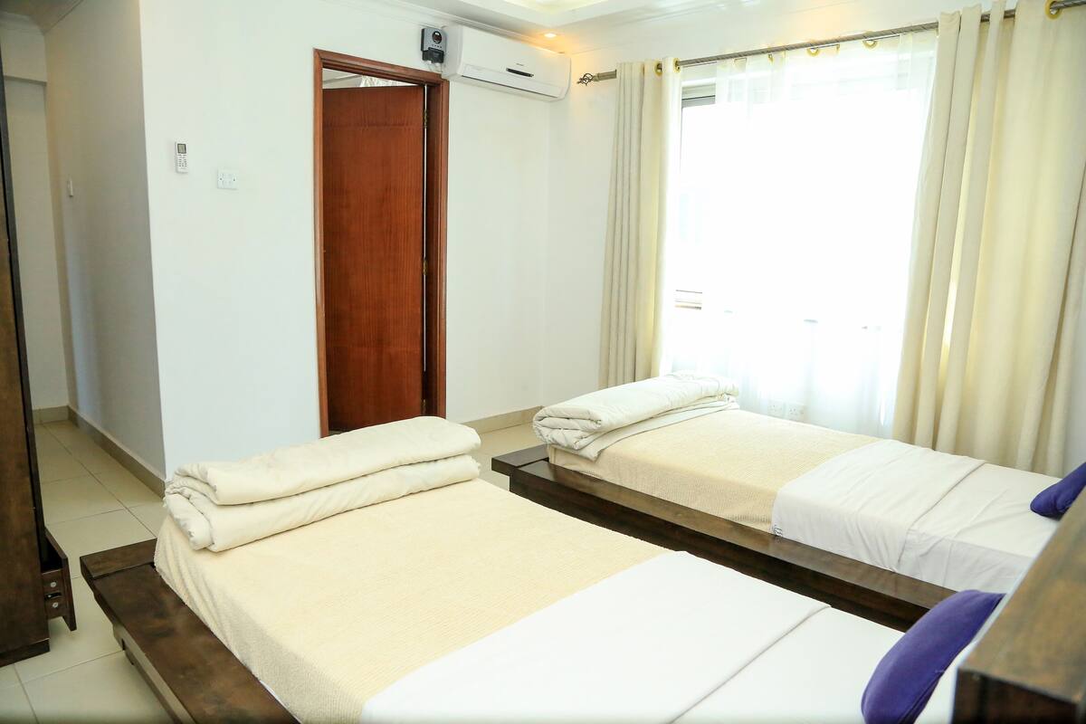 A hotel room with two beds, a wooden door, an air conditioning unit, and a window with curtains.