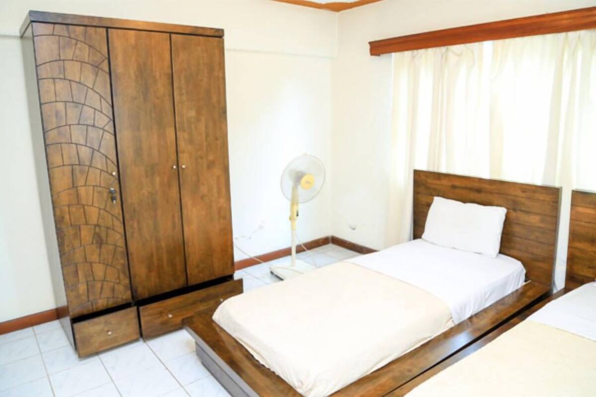 A bedroom with a wooden wardrobe, a bed with white bedding, a bedside table, and a standing fan.