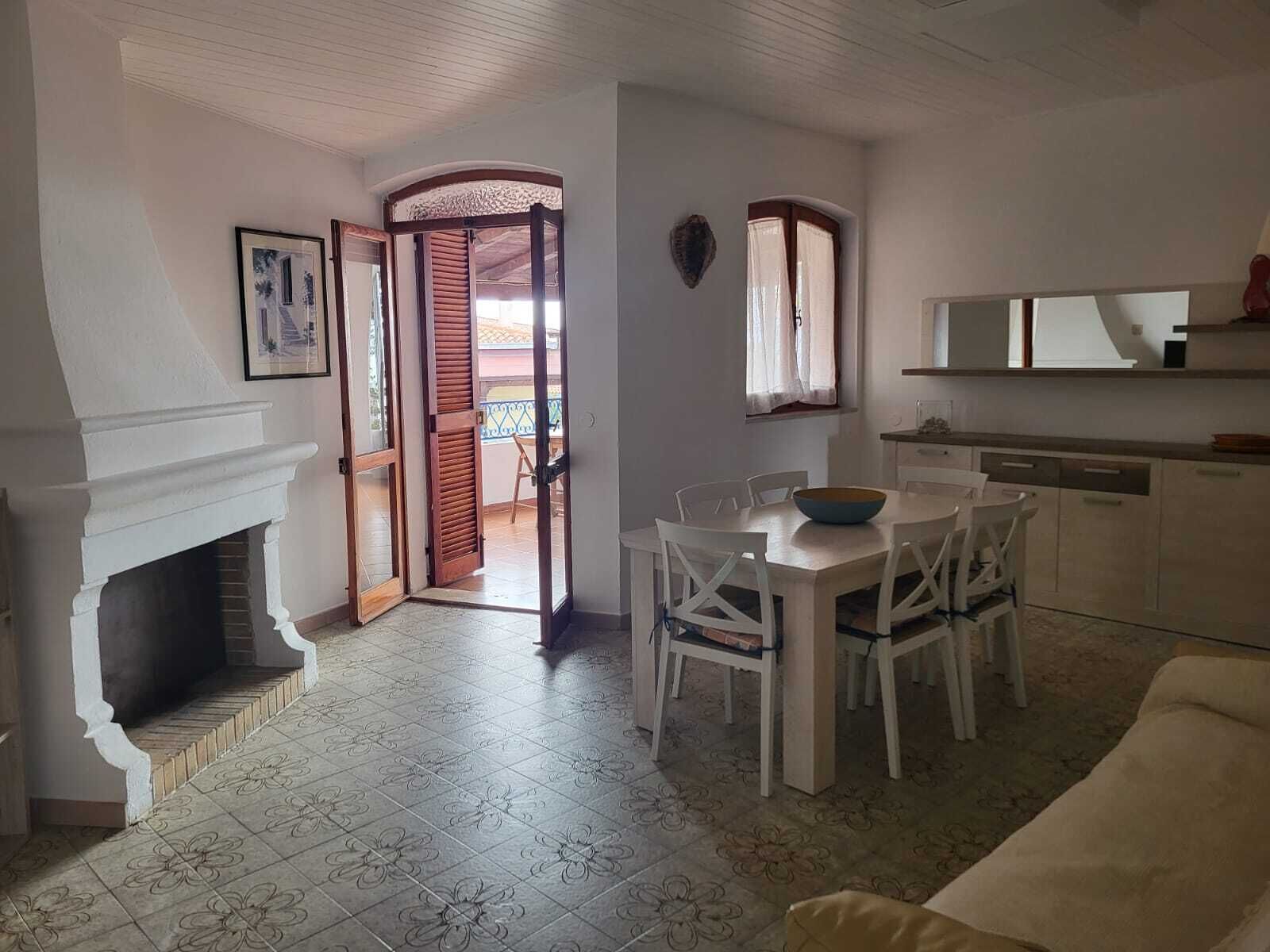 Zona de menjador amb taula i cadires blanques, una llar de foc i vista a un balcó per una porta oberta.