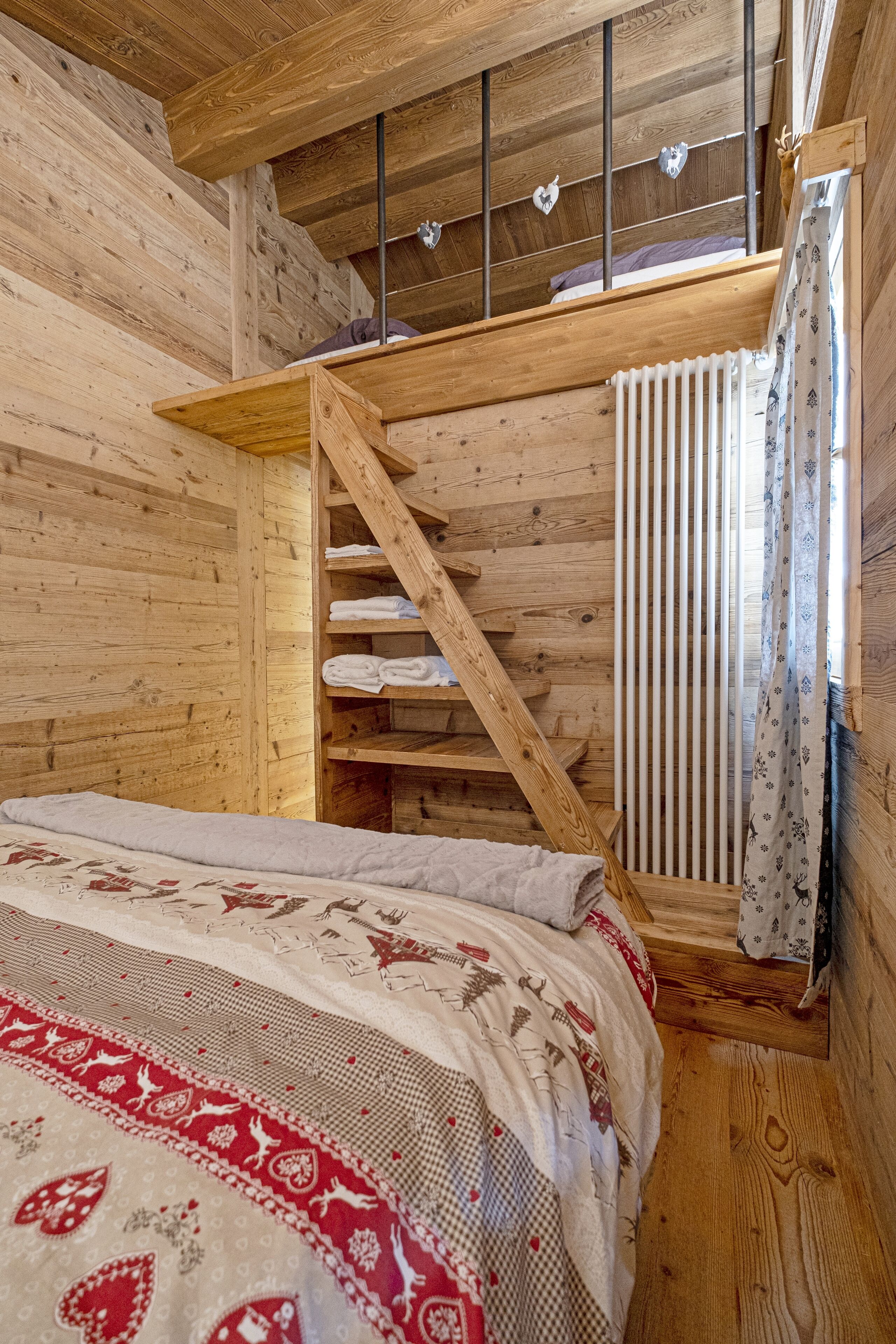 A wooden cabin room with a bed, a wooden staircase, and a window with curtains.