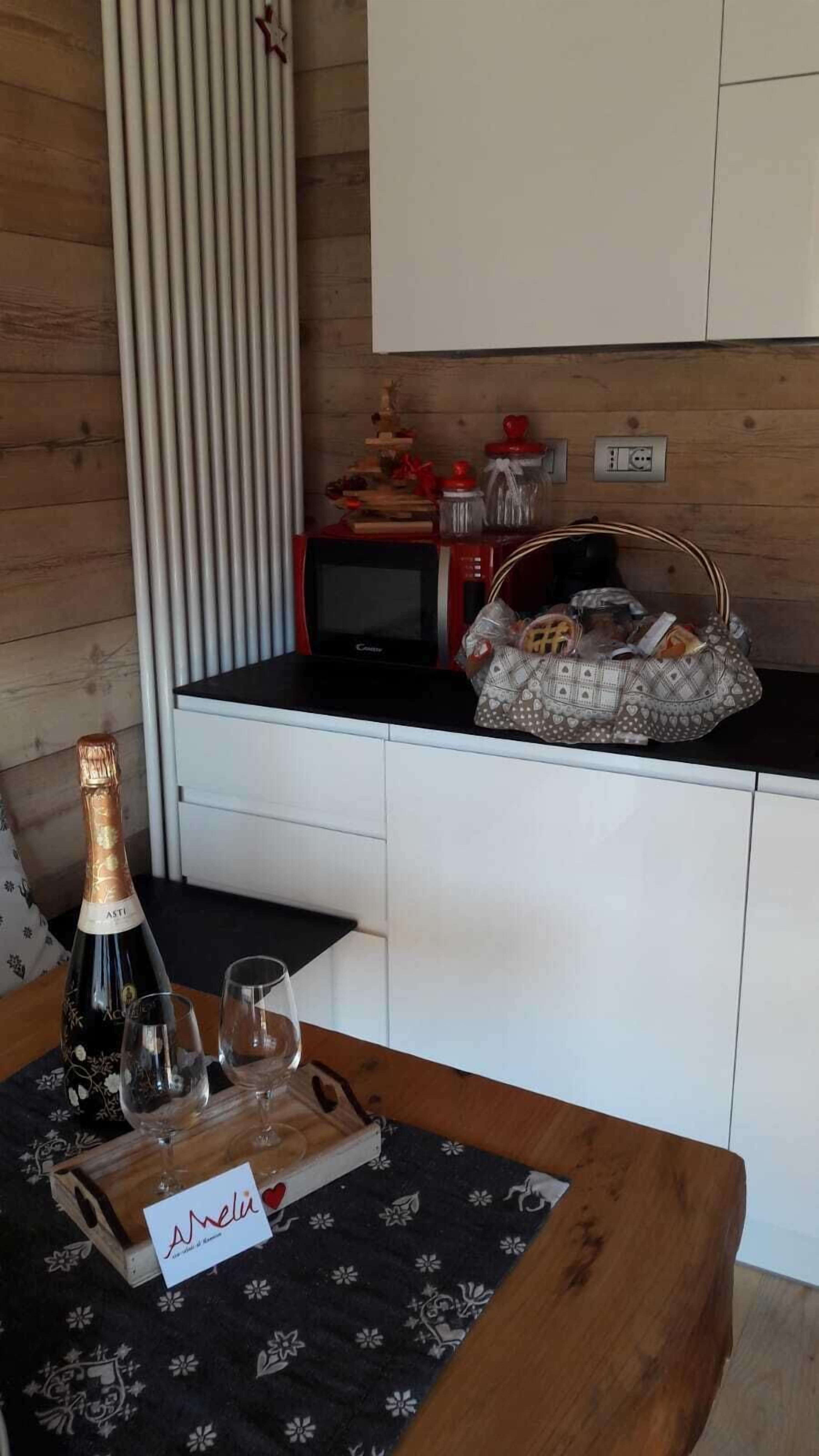 Una cocina con encimera de madera, un microondas, una cesta con utensilios de cocina, una botella de champán y dos copas sobre una mesa.