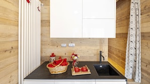A compact kitchen with wooden walls, a black countertop, a white cabinet, a sink, and a decorative basket with red items. - Amelù (Ostana)