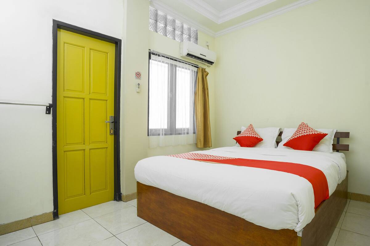 A room with a yellow door, a bed with white bedding and red accents, a window with curtains, and an air conditioning unit.