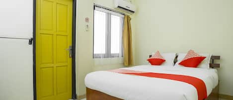 A room with a yellow door, a bed with white bedding and red accents, a window with curtains, and an air conditioning unit.