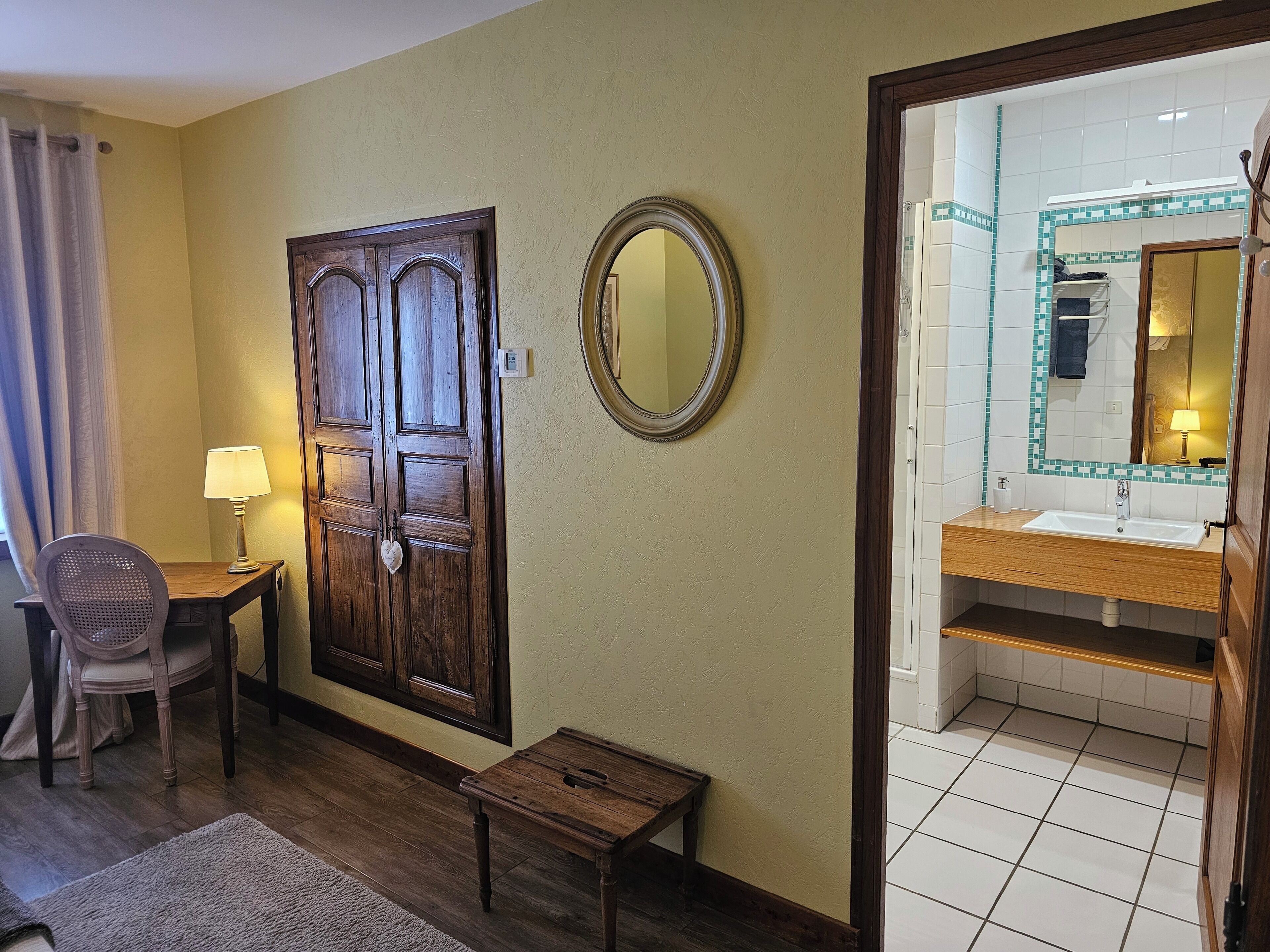 A hotel room with a wooden door, a small table, a chair, a lamp, a mirror, and a bathroom with a sink and mirror.