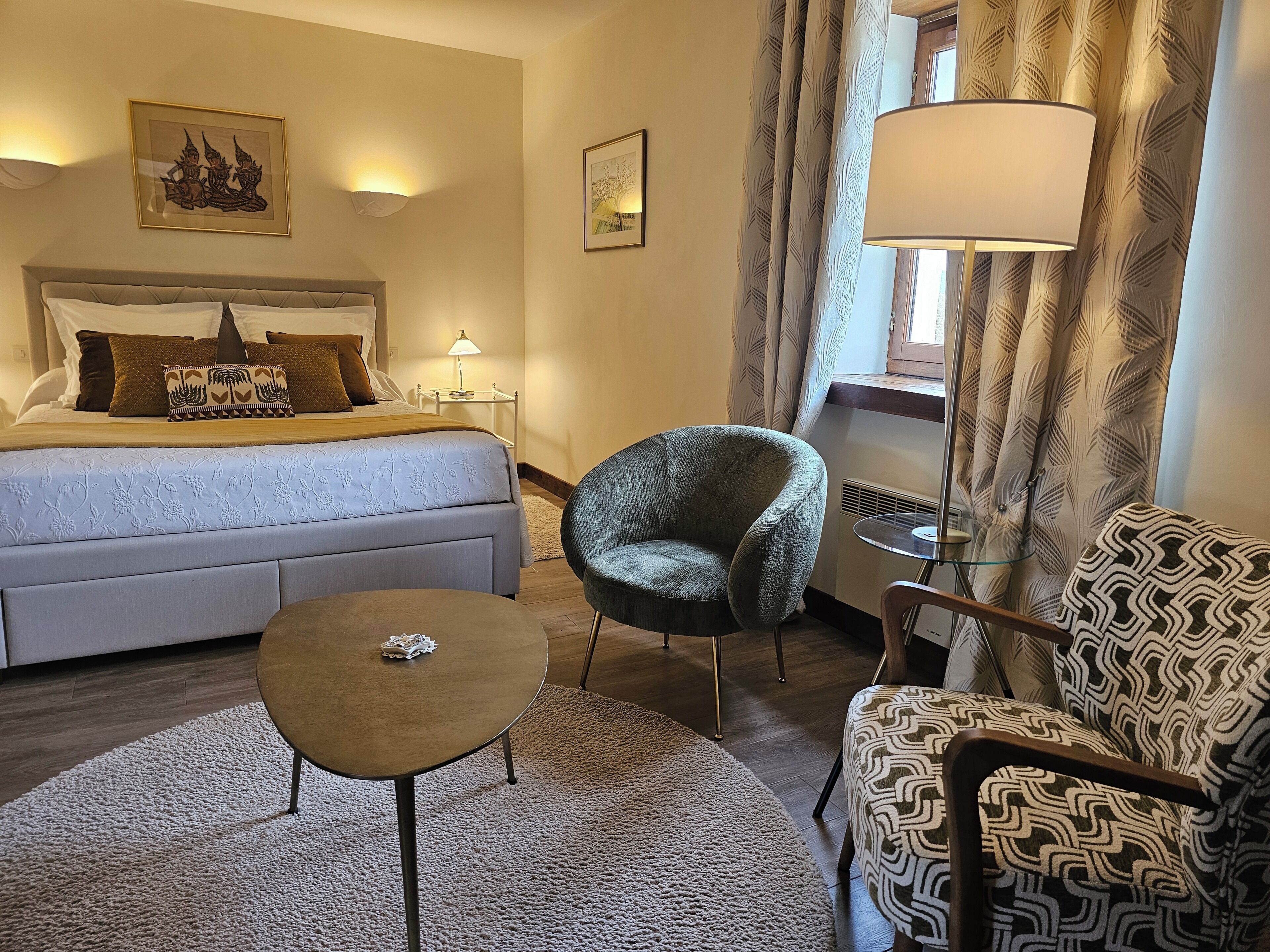 A hotel room with a bed, a chair, a small table, a lamp, and curtains.