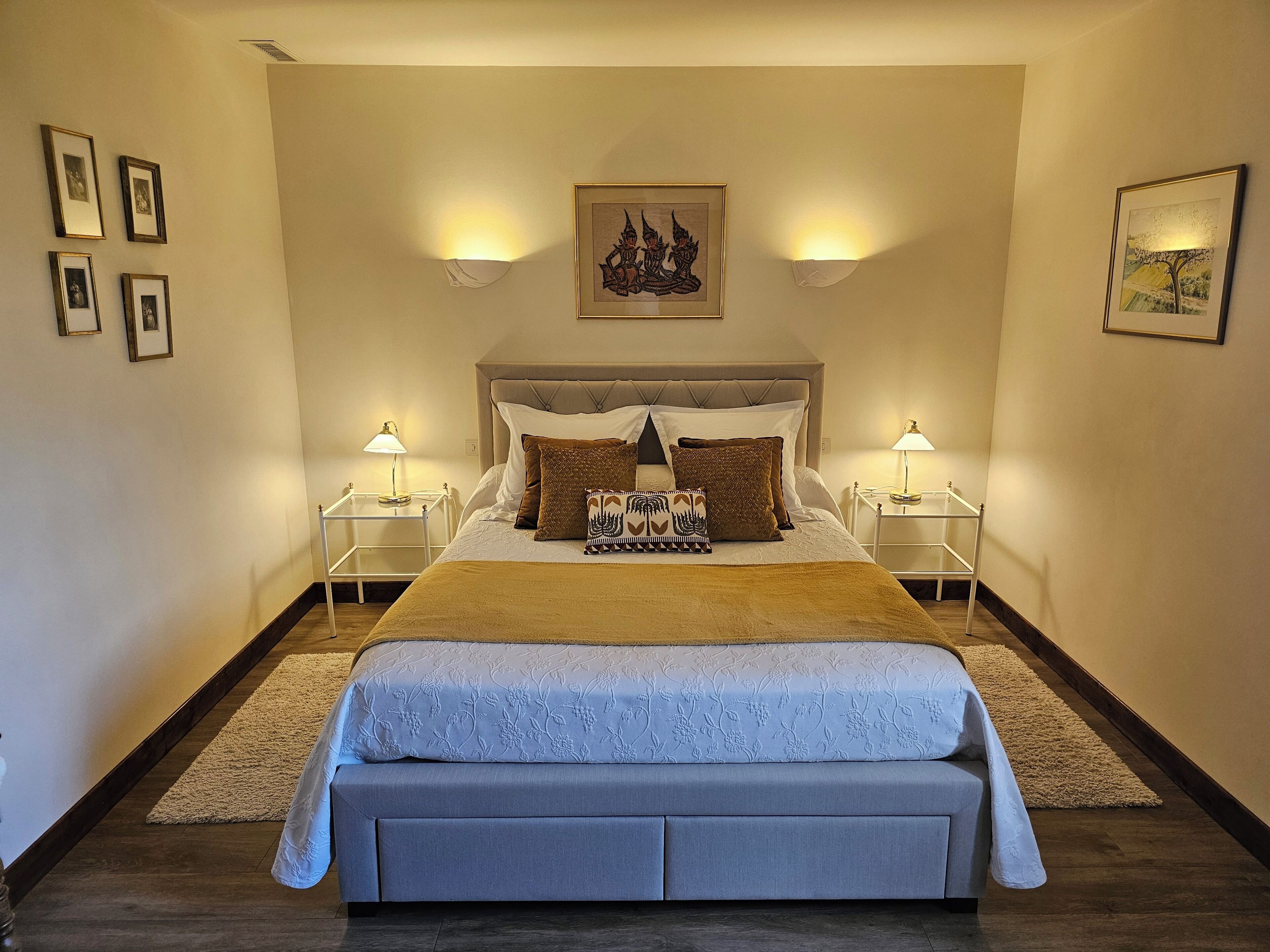 Une chambre à coucher avec un grand lit, deux tables de chevet avec des lampes, une tête de lit et des œuvres d’art encadrées accrochées aux murs.