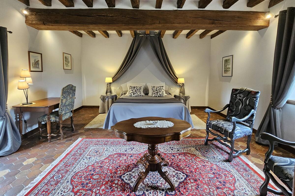 A bedroom with a large bed, a wooden table, two chairs, a patterned rug, and exposed wooden beams on the ceiling.