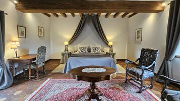 A bedroom with a large bed, a wooden table, two chairs, a patterned rug, and exposed wooden beams on the ceiling.