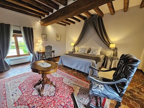 A bedroom with a bed, a chair, a table, a window with curtains, and a radiator.