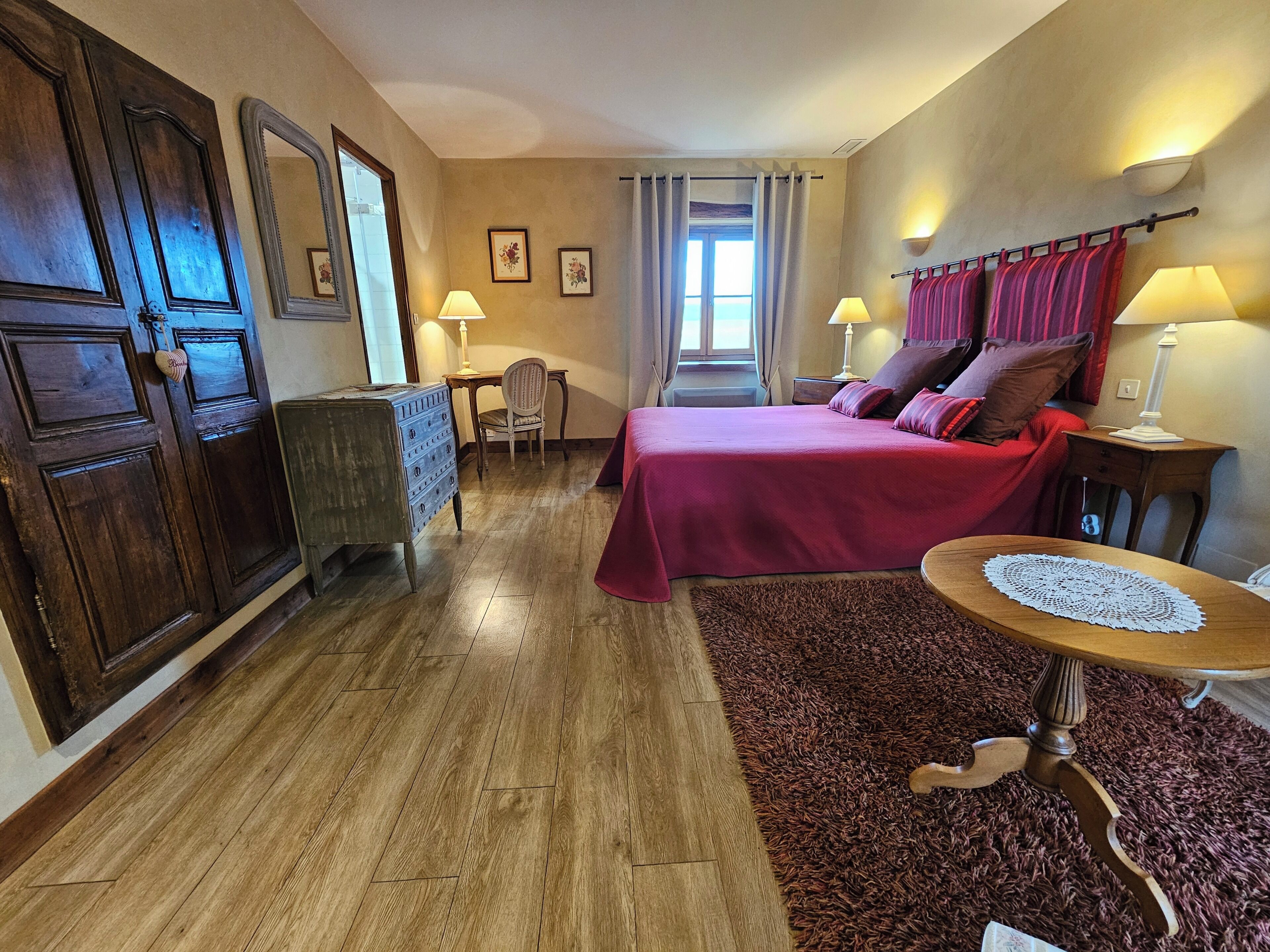 A hotel room with a wooden floor, a bed with red bedding, a wooden dresser, a chair, a small table with a doily, and a window with curtains.
