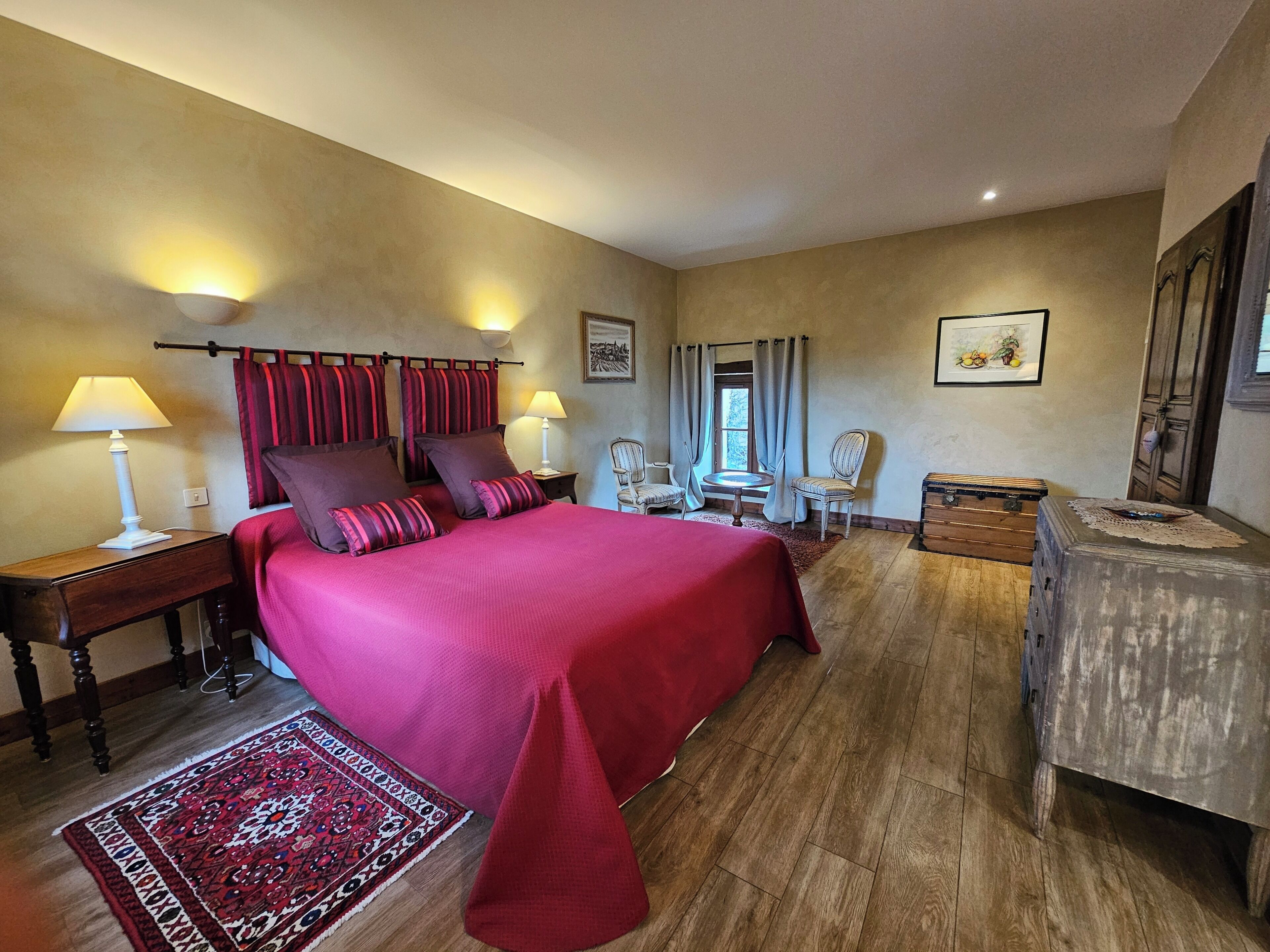 Une chambre d’hôtel avec un grand lit, deux tables de chevet, une petite table ronde, deux chaises, une commode en bois et une fenêtre avec des rideaux.