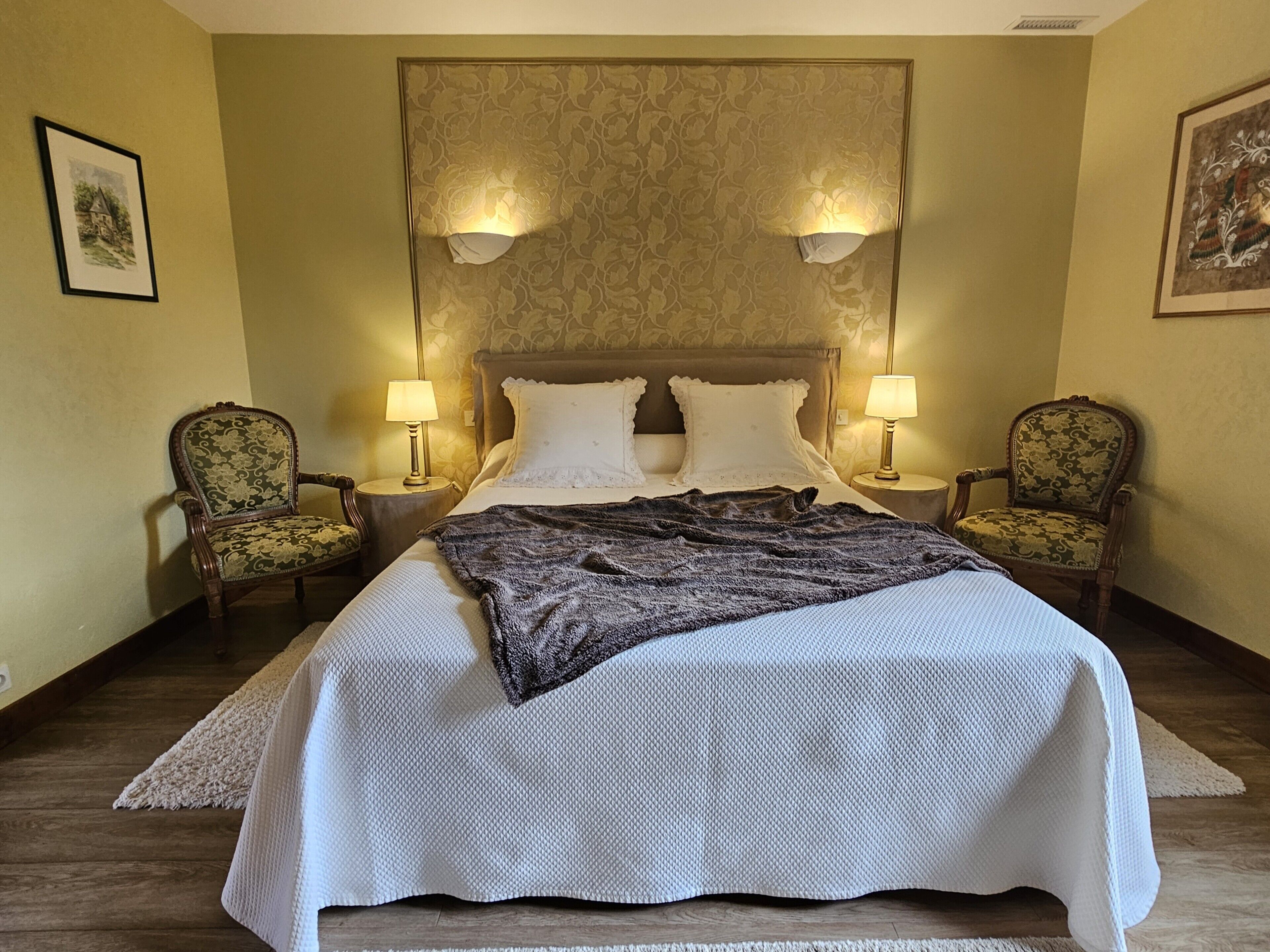 A neatly made bed with a white and gray blanket, two lamps on nightstands, and two upholstered chairs with floral upholstery.