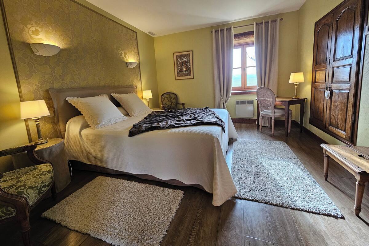 A hotel room with a bed, two bedside tables with lamps, a desk with a chair, a large wardrobe, and a window with curtains.