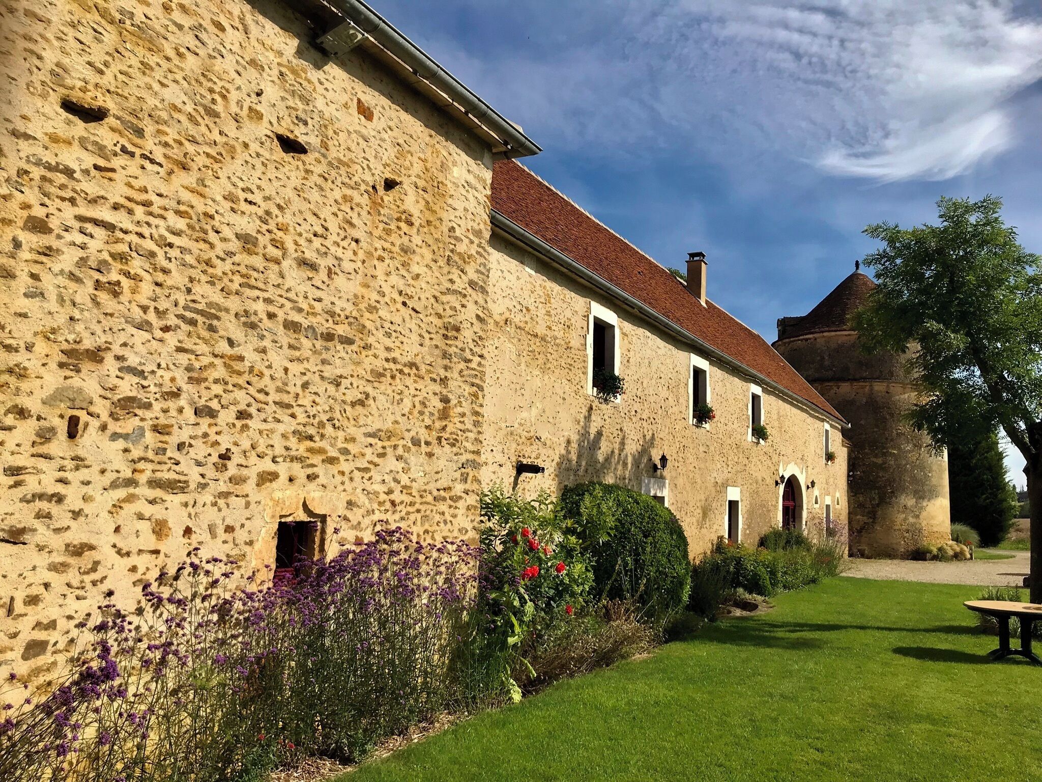 A stone building with a red-tiled roof, a garden with purple flowers, and a clear sky.